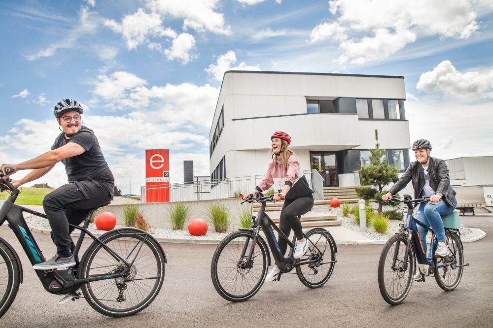 3 Mitarbeiter:innen der e2 elektro GmbH radeln mit 3 E-Bikes vor dem Firmengebäude.